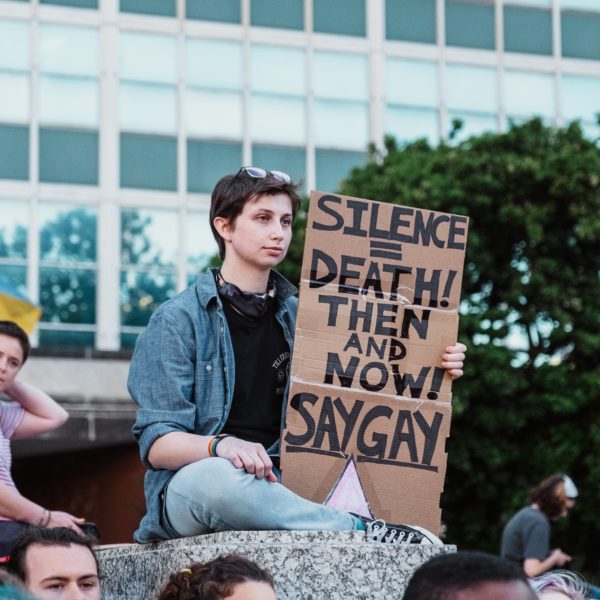 someone sitting above a crowd holding a sign that says silence = death then and now say gay